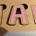Extreme macro of a single pink-painted letter A showing wood grain visible through the thin matte paint and the laser-charred edge line