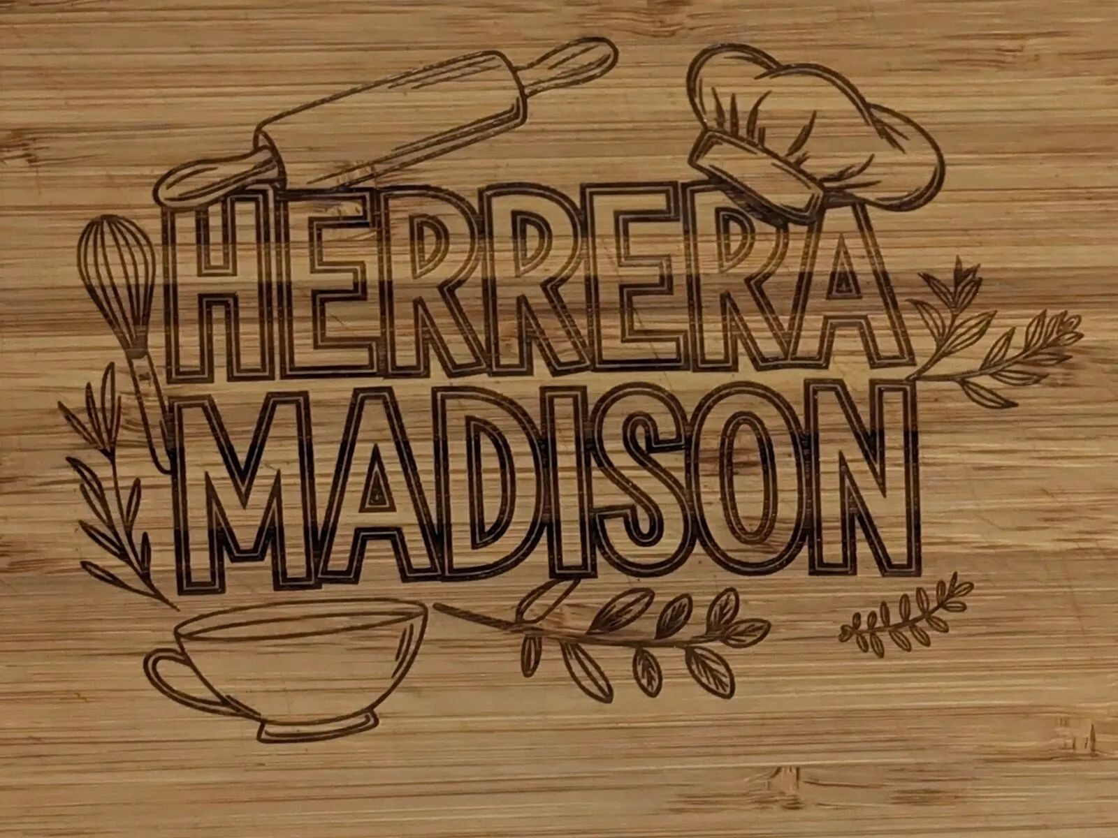 Close view of two family names engraved into a bamboo cutting board, surrounded by engraved baking tools and leaves