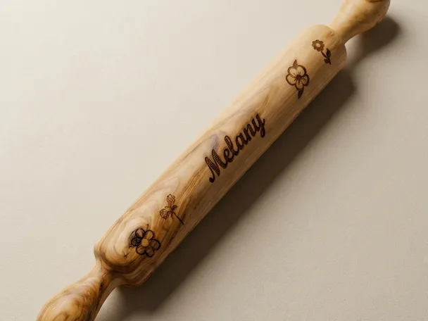Solid wood rolling pin resting on a warm neutral surface, turned handles at both ends and a laser-engraved script name across the barrel with small flower motifs on either side