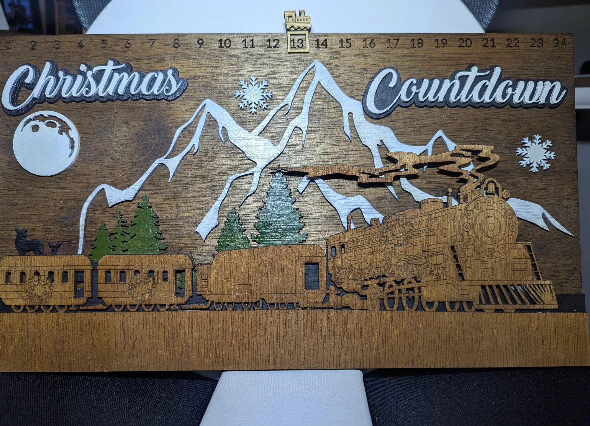 Layered wood Christmas countdown sign: a dark walnut backer with days 1 to 24 engraved along the top edge, white painted mountains and moon, green pine trees, a standing deer, and a golden steam train with a smoke plume, standing on a full-width base ledge