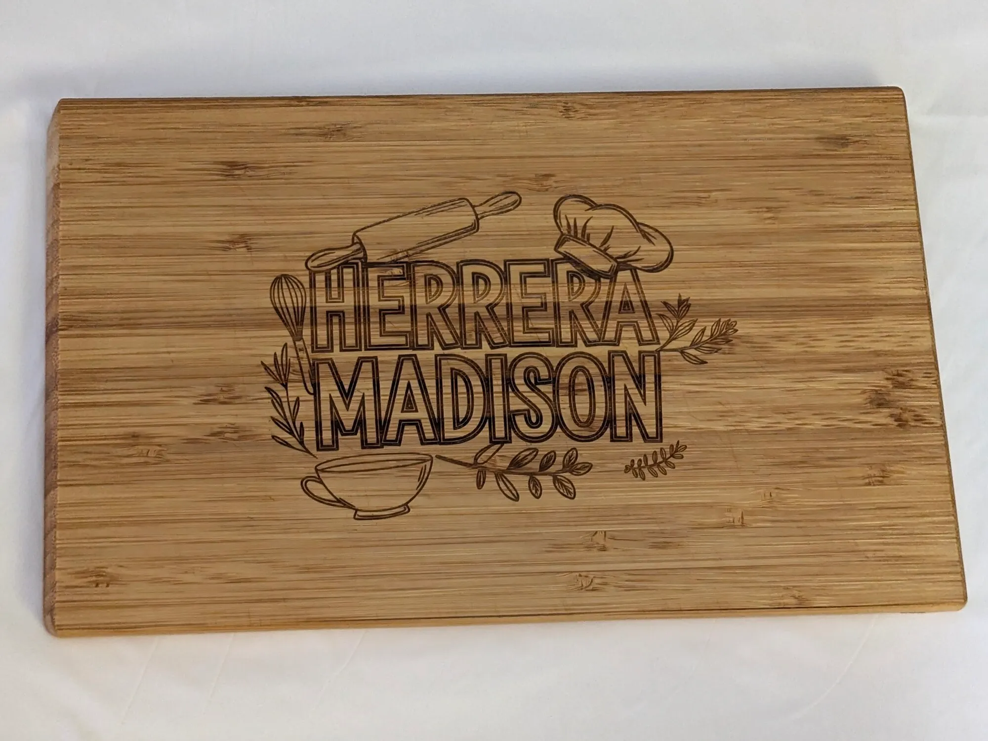 Bamboo cutting board engraved with a family name in outlined block capitals inside a wreath of kitchen line art