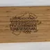 Bamboo cutting board on a white cloth, engraved with the family name Herrera Madison in outlined block capitals inside a wreath of kitchen line art